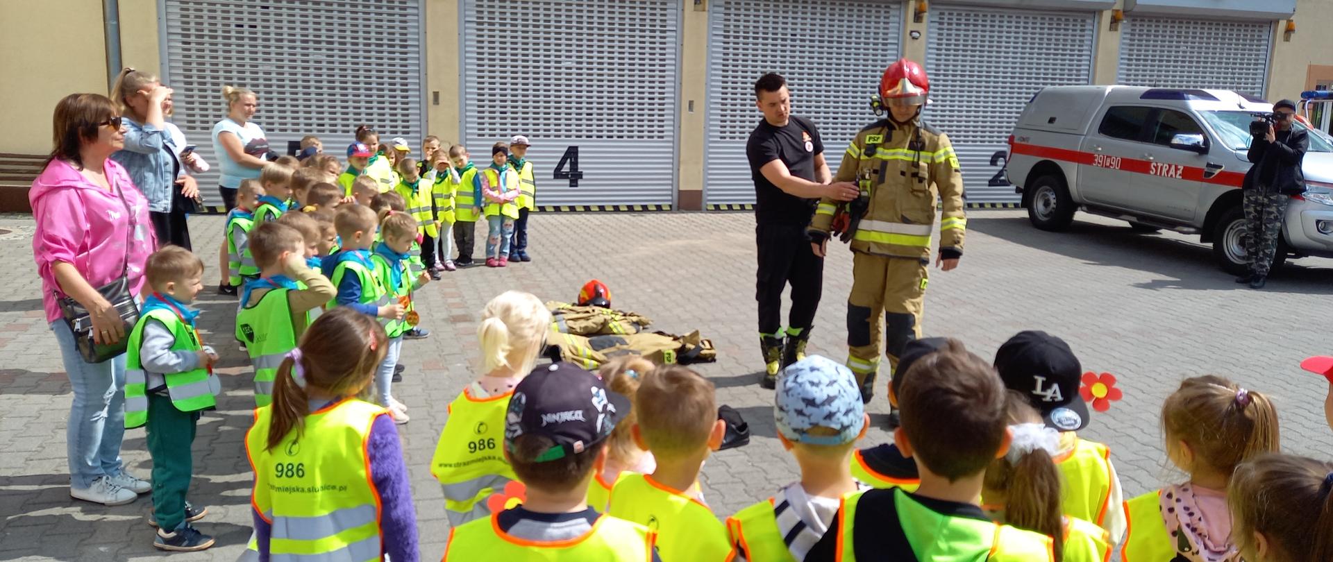 Duża grupa dzieci stojąca na placu wewnętrznym KP PSP w Słubicach. Przed nimi jeden strażak w umundurowaniu bojowym, kolejny prezentujący to umundurowanie.