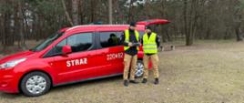 Na zdjęciu dwaj strażacy na tle samochodu czerwonego z napisem straż na drzwiach przednich i 220T82 na drzwiach tylnych, w lesie, trzymają w ręku tablet do sterowania dronem. Strażacy w mundurach i kamizelkach odblaskowych.