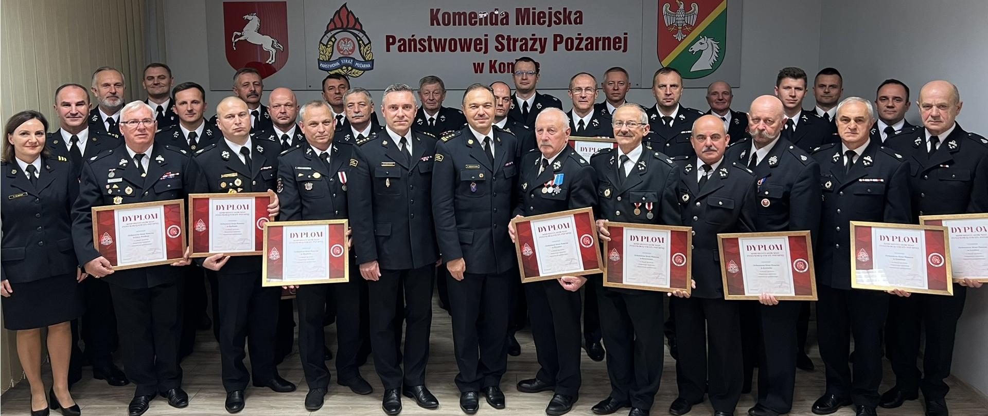 Druhowie OSP oraz strażacy stoją w świetlicy Komendy podczas przekazania dyplomów Komendanta Głównego Państwowej Straży Pożarnej z okazji 30-lecia Krajowego Systemu Ratowniczo-Gaśniczego. Druhowie trzymają w rękach dyplomy