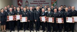 Druhowie OSP oraz strażacy stoją w świetlicy Komendy podczas przekazania dyplomów Komendanta Głównego Państwowej Straży Pożarnej z okazji 30-lecia Krajowego Systemu Ratowniczo-Gaśniczego. Druhowie trzymają w rękach dyplomy