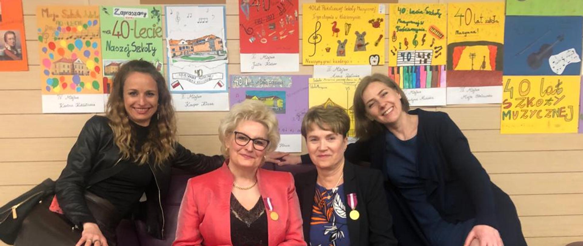 Na zdjęciu cztery panie na śliwkowej sofie. Pani z prawej i lewej strony ubrane w ciemnych kolorach, głównie czerń. Panie po środku: jedna w okularach i czerwonej marynarce, a druga granatowa sukienka i czarna marynarka. Panie po środku mają złote medale za długoletnią służbę. Za nimi w tle wystawa plakatów związanych z jubileuszem 40- ecia szkoły. Plakaty zawieszone są za beżowej ścianie wykonanej w desek.