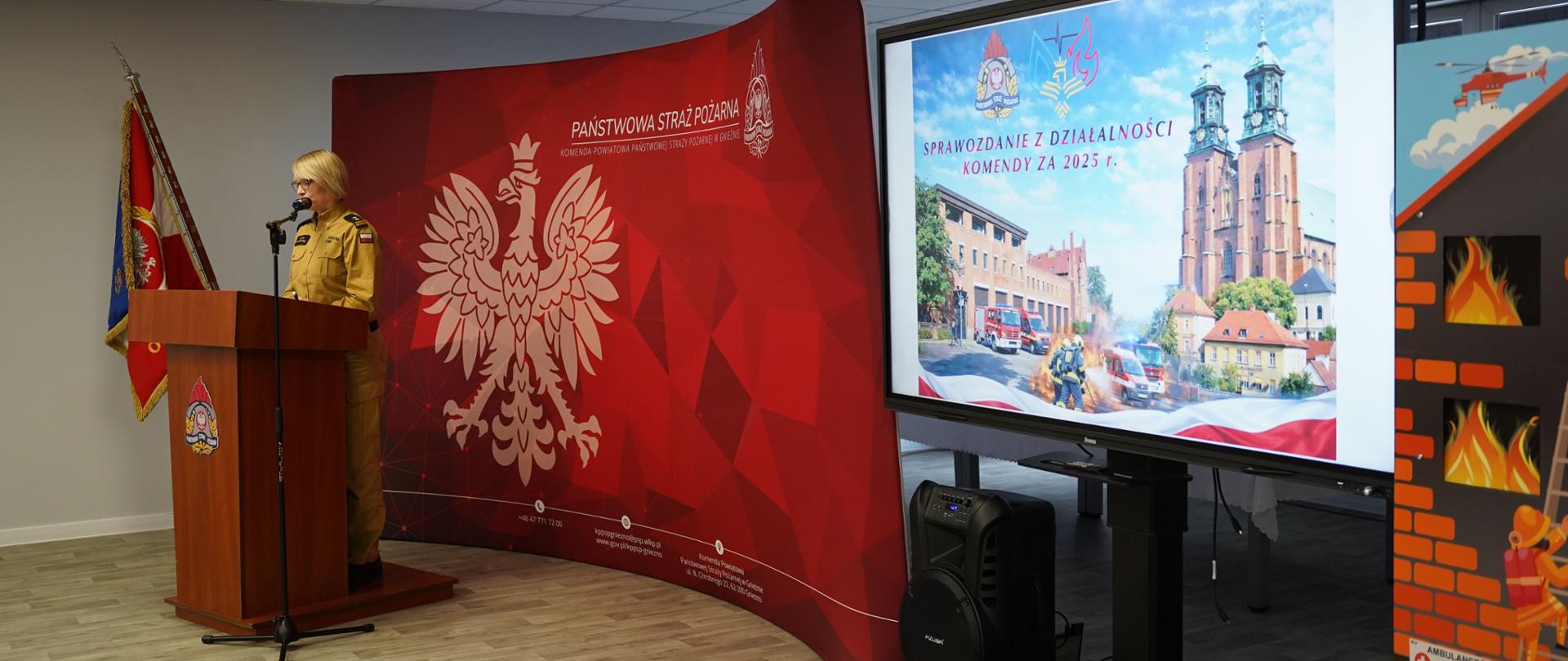 Funkcjonariuszka Państwowej Straży Pożarnej w mundurze stoi przy mównicy i wygłasza przemówienie podczas prezentacji. Obok widoczny jest ekran z napisem „Sprawozdanie z działalności komendy za 2025 r.” oraz grafiką przedstawiającą wozy strażackie i zabytkowy kościół, a w tle czerwone tło z godłem PSP