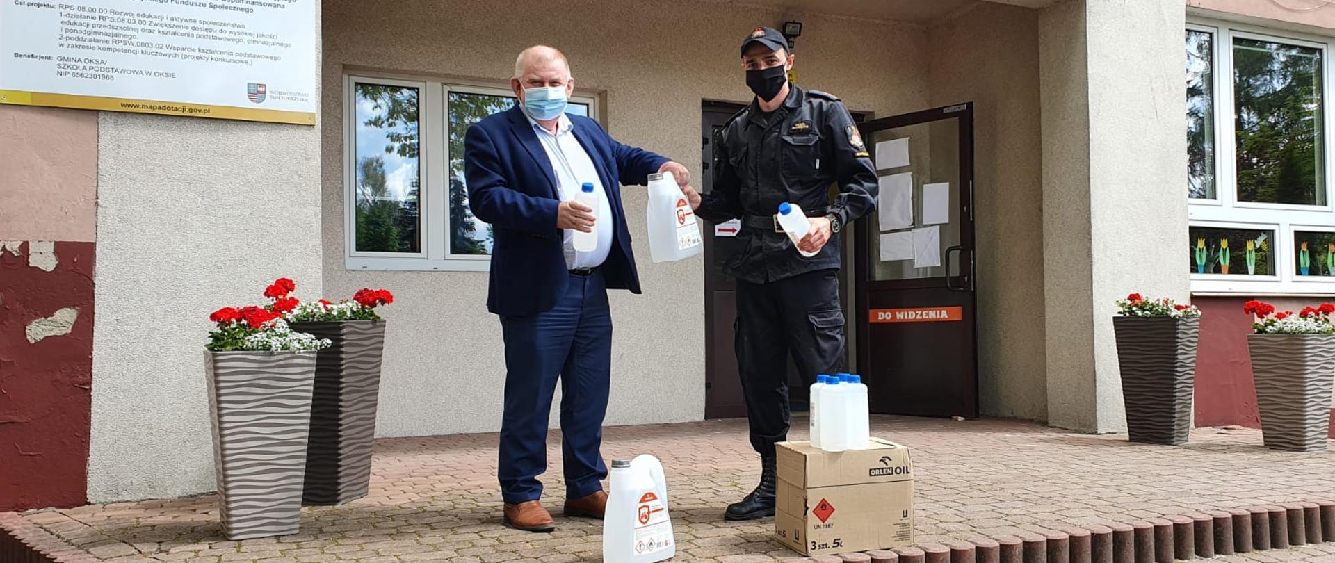 Zdjęcie obrazuje strażaka w stroju dowódczo-sztabowym, który przekazuje środki ochrony indywidualnej dyrektorowi placówki oświatowej. Zdjęcie zrobione przed budynkiem szkoły. Obaj mężczyźnie posiadają maseczki ochronne zakrywające usta i nos. 