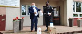 Zdjęcie obrazuje strażaka w stroju dowódczo-sztabowym, który przekazuje środki ochrony indywidualnej dyrektorowi placówki oświatowej. Zdjęcie zrobione przed budynkiem szkoły. Obaj mężczyźnie posiadają maseczki ochronne zakrywające usta i nos.