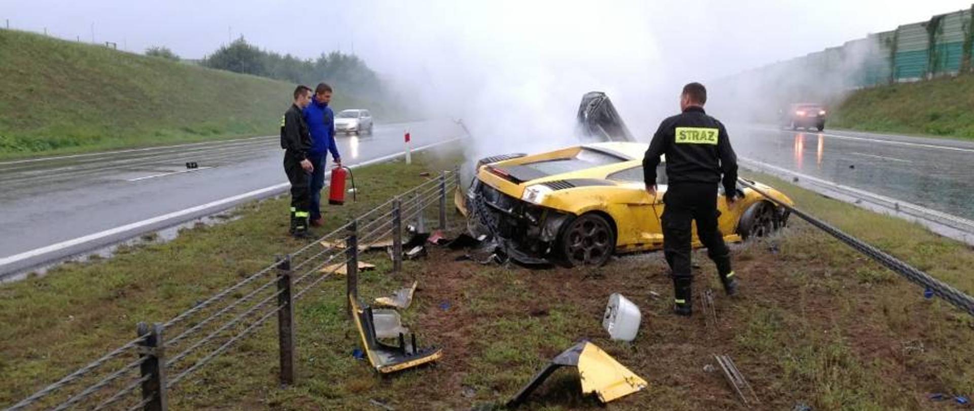 Zdjęcie przedstawia trzy osoby, dwie z nich w mundurach strażackich, jedna ubrana po cywilnemu trzyma gaśnicę w ręku przy dymiącym się żółtym samochodzie marki Lamborghini i jego kawałkach rozrzuconych na poboczu autostrady A1.
