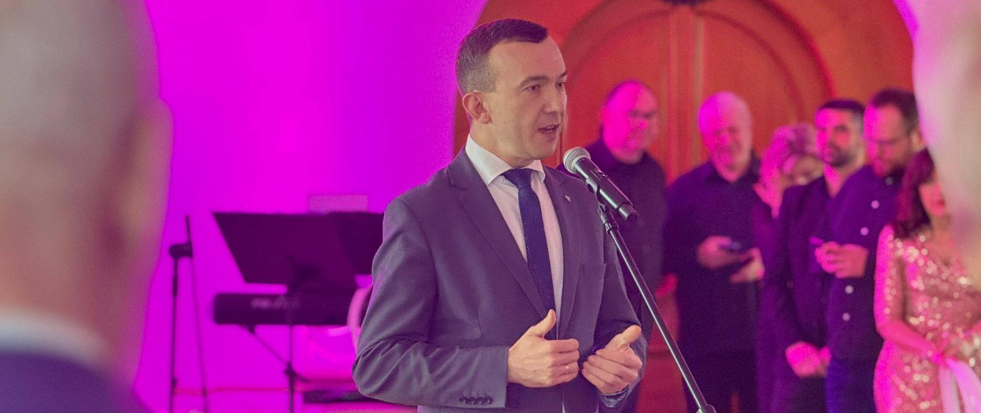 Wicewojewoda Mateusz Szauer przemawia gestykulując rękoma, jest ubrany w szary garnitur 