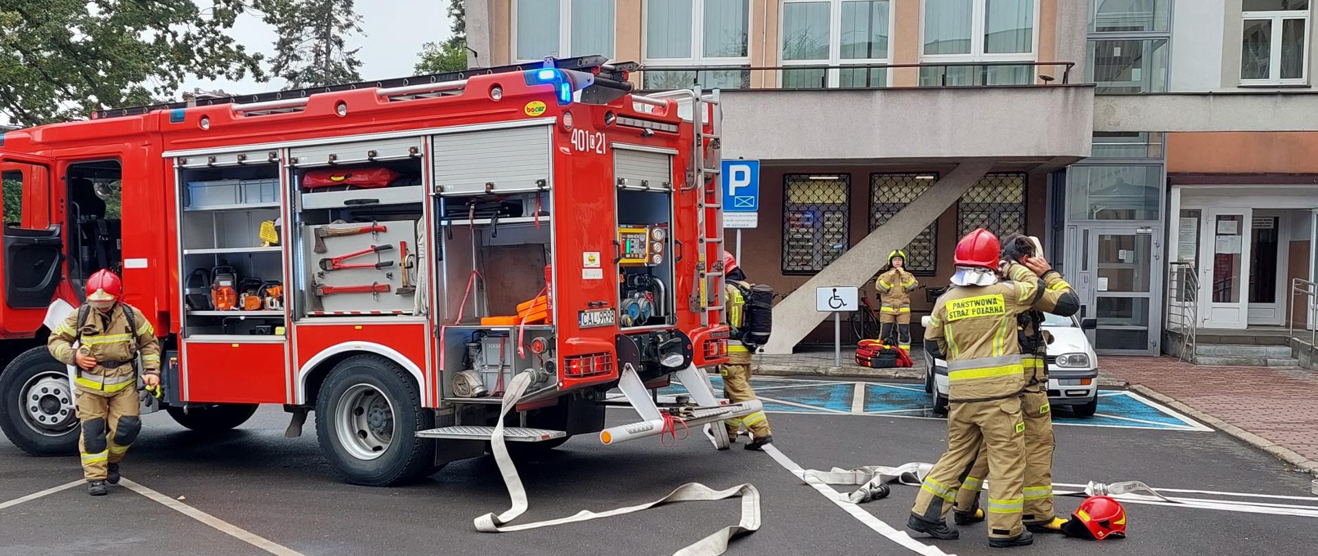 W lewej części zdjęcia na wyasfaltowanym placu stoi czerwony ciężarowy samochód strażacki z otwartymi skrytkami, w których znajduje się sprzęt pożarniczy. Przed pojazdem oraz po prawej stronie widać ubranych na żółto strażaków przygotowujących się do prowadzenia działań gaśniczych. Między nimi rozwinięte są jasne węże strażackie. Tło zdjęcia stanowi jasna ściana budynku z licznymi przeszkleniami.