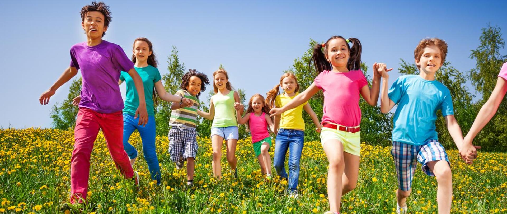 grafika kolorowa, zielona łąka i żółte kwiatki mlecze po której biegają chłopcy i dziewczynki ubrani w kolorowe stroje