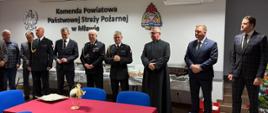 Grupa mężczyzn stoi w sali konferencyjnej Komendy Powiatowej Państwowej Straży Pożarnej w Mławie. Część z nich jest w mundurach strażackich, inni w garniturach. Na ścianie widać godło państwowe oraz emblemat PSP. Uczestnicy stoją w rzędzie, z powagą, jak podczas oficjalnej uroczystości.
