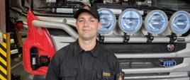 Zdjęcie przedstawia funkcjonariusza Państwowej Straży Pożarnej w Ostrzeszowie na tle pojazdu gaśniczego. Zdjęcie wykonane w garażu w porze dziennej.