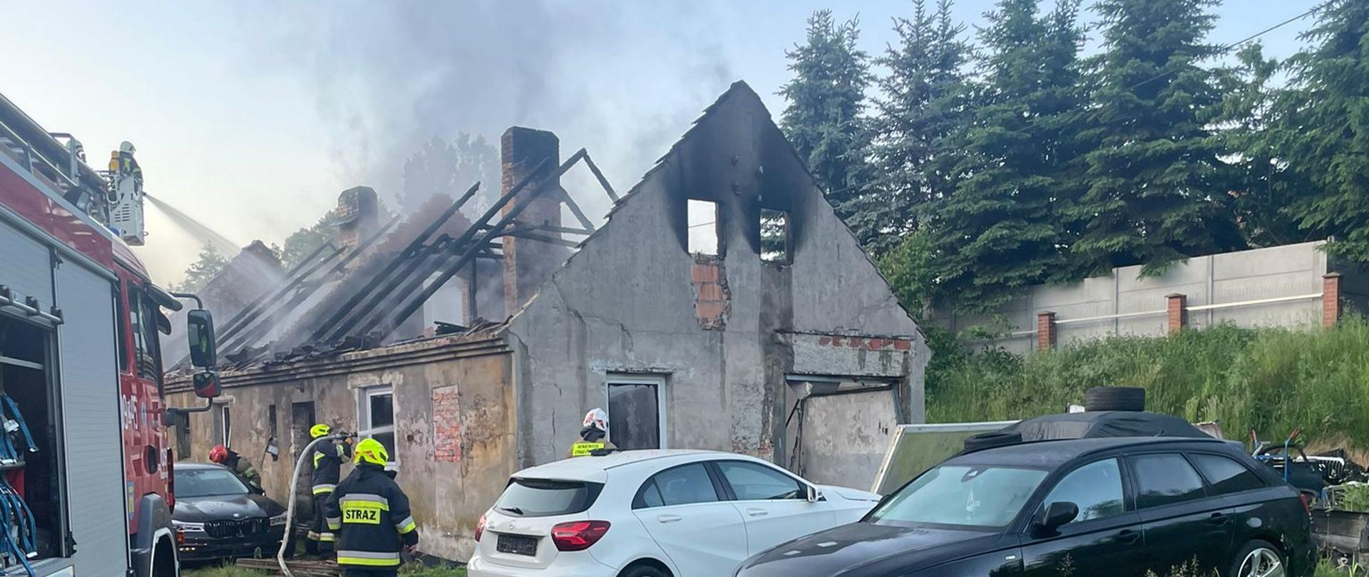 Dzień. W tle parterowy budynek gospodarczy z całkowicie spalonym dachem. Pozostało kilka nadpalonych belek. Z budynku unosi się biały dym. Z lewej strony widać cześć pojazdu ratowniczo-gaśniczego od którego biegnie wąż strażacki w stronę budynku. Przy budynku stoi trzech strażaków ubranych w ubrania specjalne w kolorze granatowym. Przy budynku stoją dwa samochody osobowe.