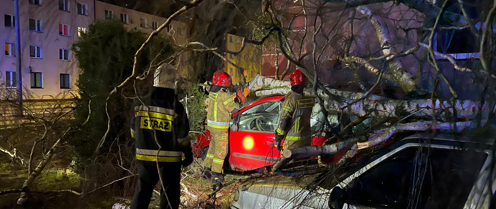Zdjęcie przedstawia trzech strażaków, którzy w porze nocnej usuwają powalone drzewo na dwa samochody osobowe stojące na parkingu. W tle widoczne budynki mieszkalne wielorodzinne. 