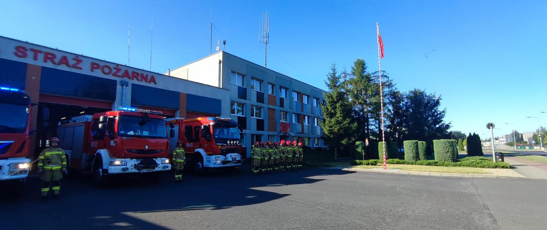 Strażacy stoją w dwuszeregu obok ustawionych pojazdów pożarniczych. W tle widoczny dudynek komendy.