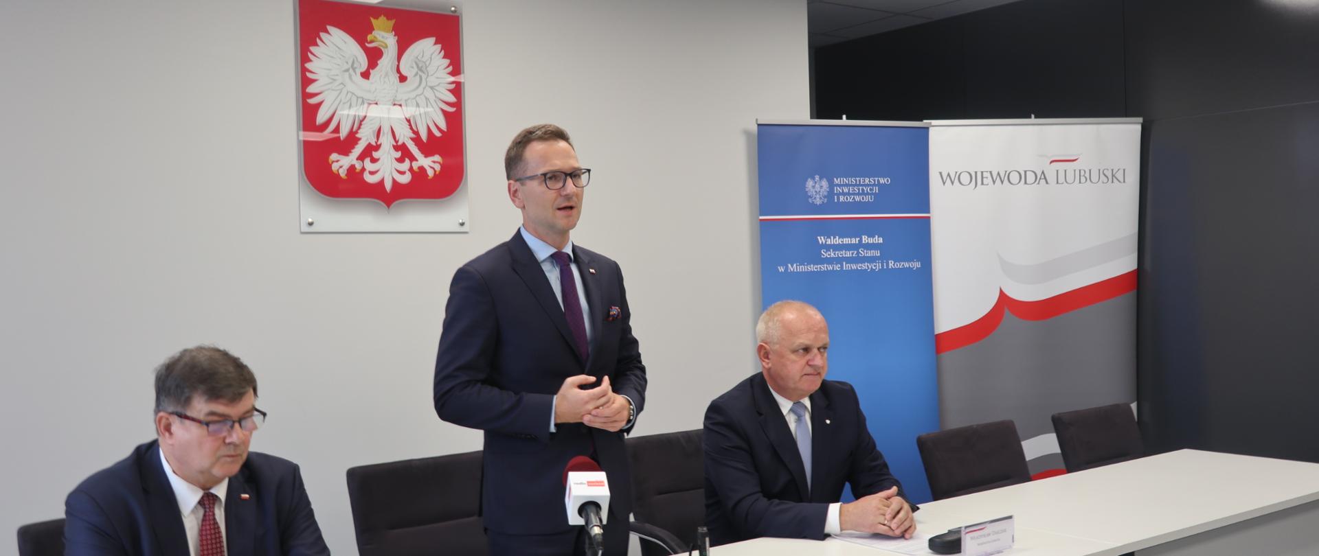 Na zdjęcie trzy osoby za stołem, w środku stoi wiceminister Waldemar Buda, w tle godło Polski.