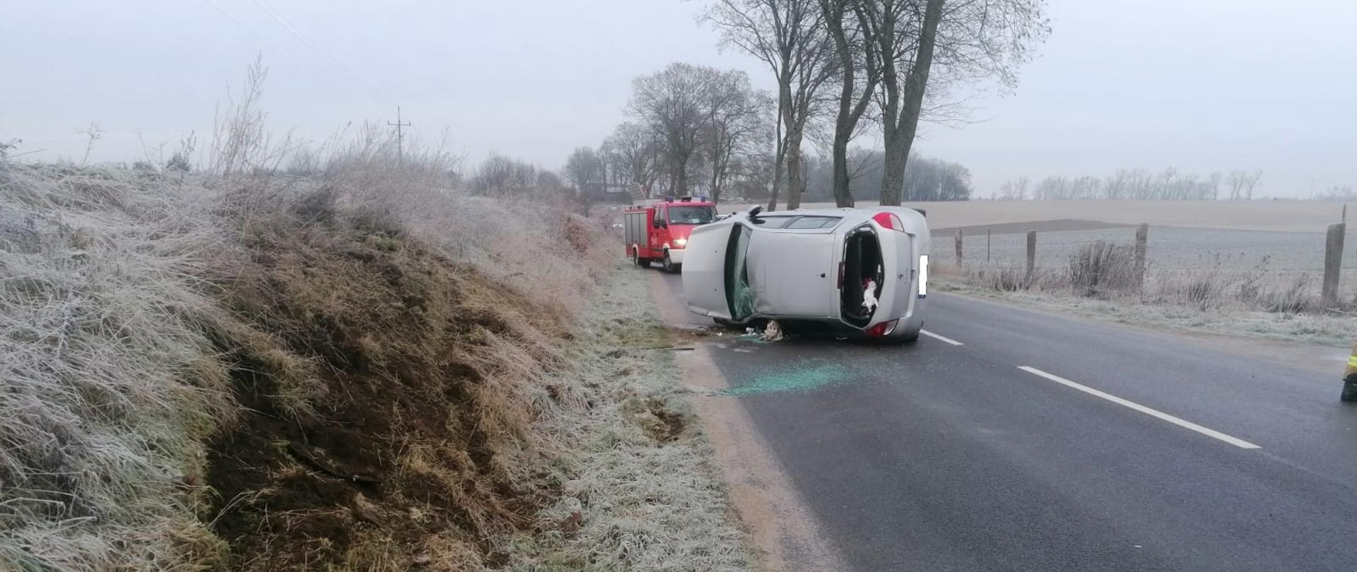 Zdjęcie przedstawia jezdnię na której na łuku drogi leży biały samochód osobowy. W tle widzimy zaszroniona trawę.