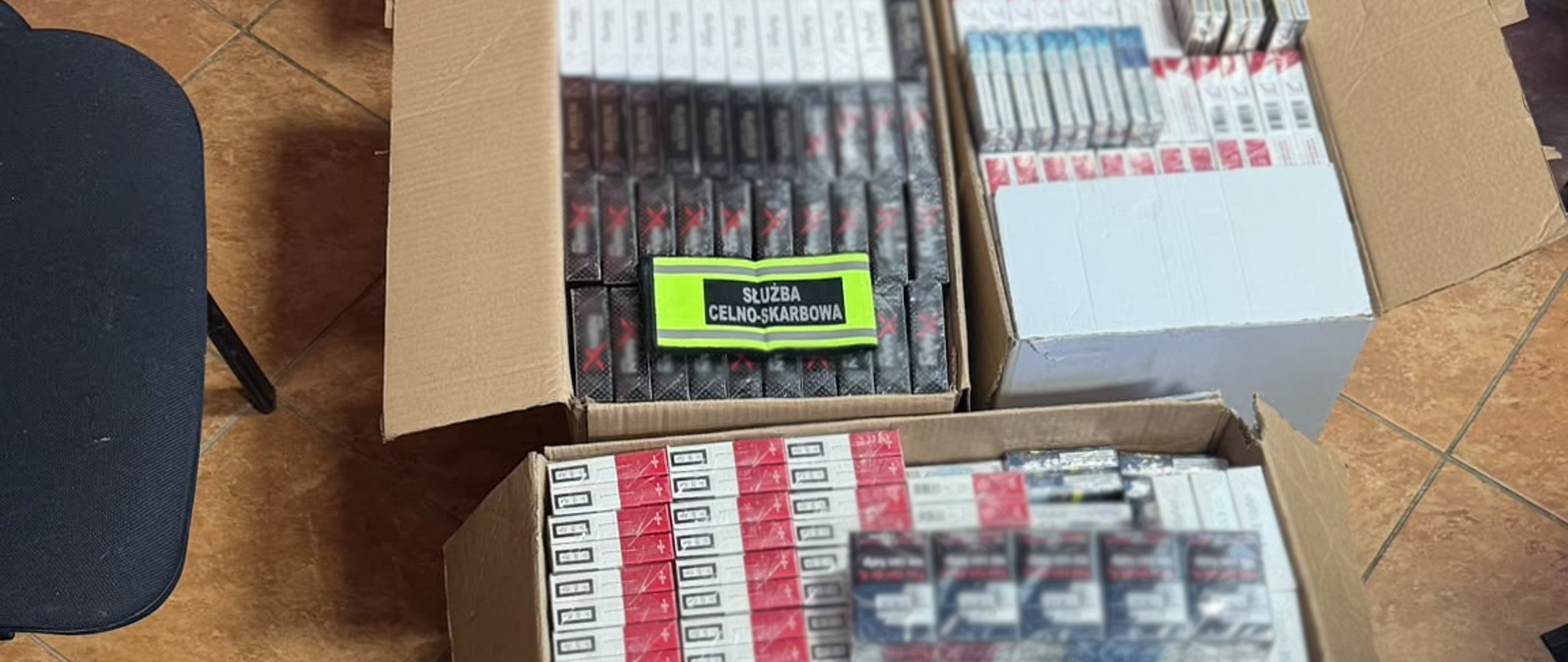 Otwarte kartony wypełnione licznymi, różnymi paczkami papierosów. Na jednej ze sztang papierosów widoczna opaska odblaskowa z czarnym napisem "SŁUŻBA CELNO-SKARBOWA".