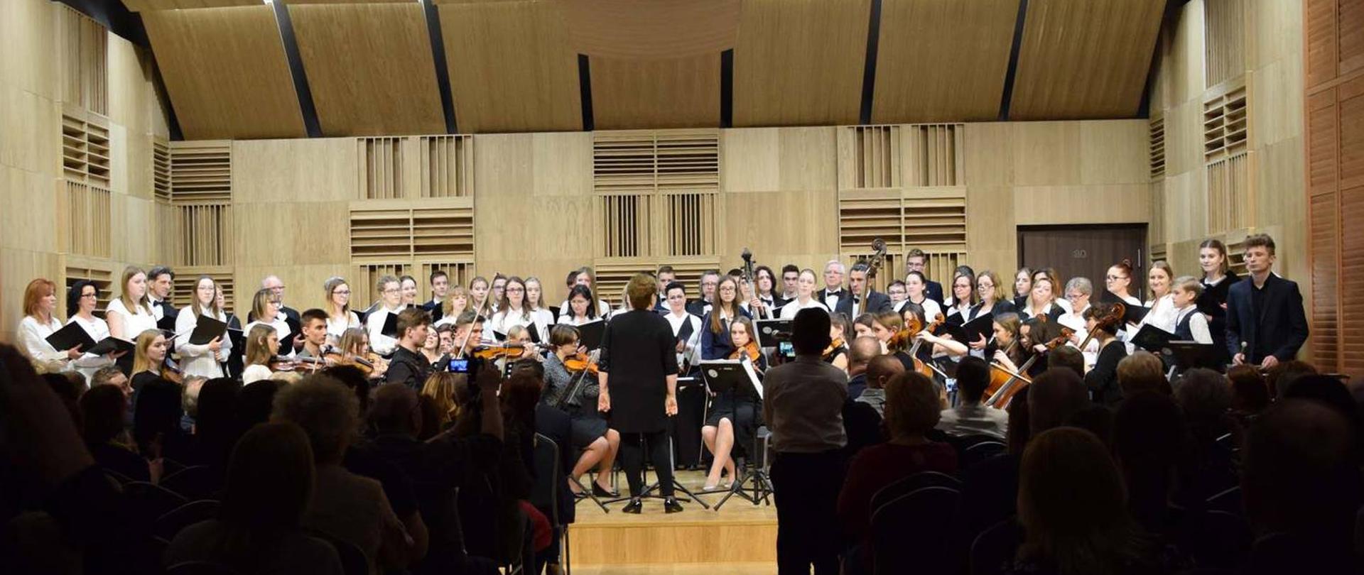 Nowoczesna sala koncertowa pełna publiczności widok od wejścia. Na estradzie duży chór i orkiestra Zespołu Szkół Muzycznych w Pile oświetlona białym światłem strefowym.