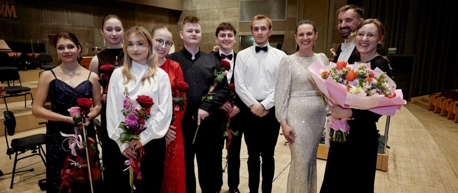 Na zdjęciu siedmiu absolwentów roku 2026 Państwowej Szkoły Muzycznej drugiego stopnia w Kaliszu wraz z dyrekcją i dyrygentem. Zdjęcie zrobione zostało na Auli UAM w Kaliszu po koncercie dyplomantów. Wszyscy ubrani są w galowe stroje a uczniowie oraz pani dyrygent trzymają w rękach kwiaty. 