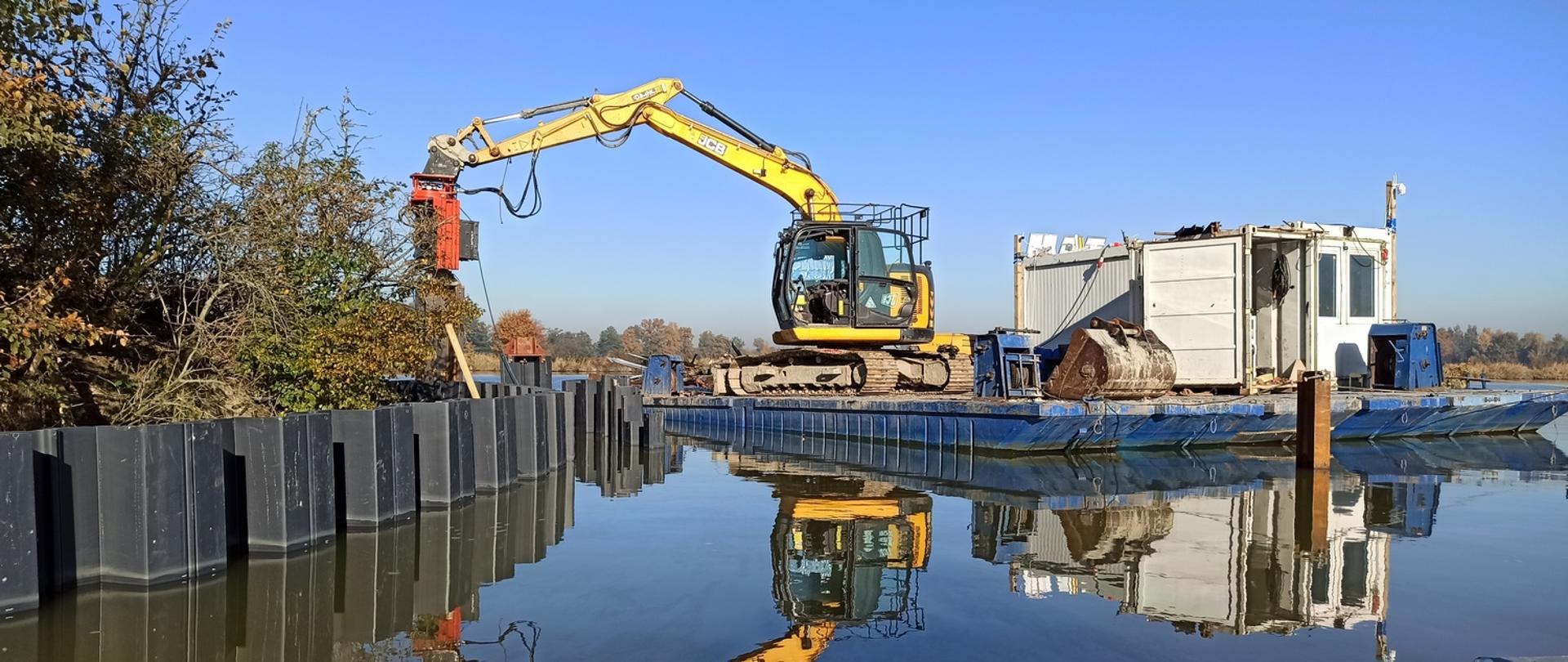 Na specjalnej platformie, tuż przy wyspie, znajduje się koparka i pozostały sprzęt budowlany.
