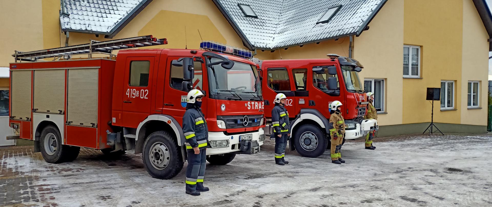 Poza zimowa, leżący śnieg na placu. Na pierwszym planie dwa stojące skosem wozy strażackie, przy autach stoją na baczność o dwóch strażaków. W tle budynek remizy. 
