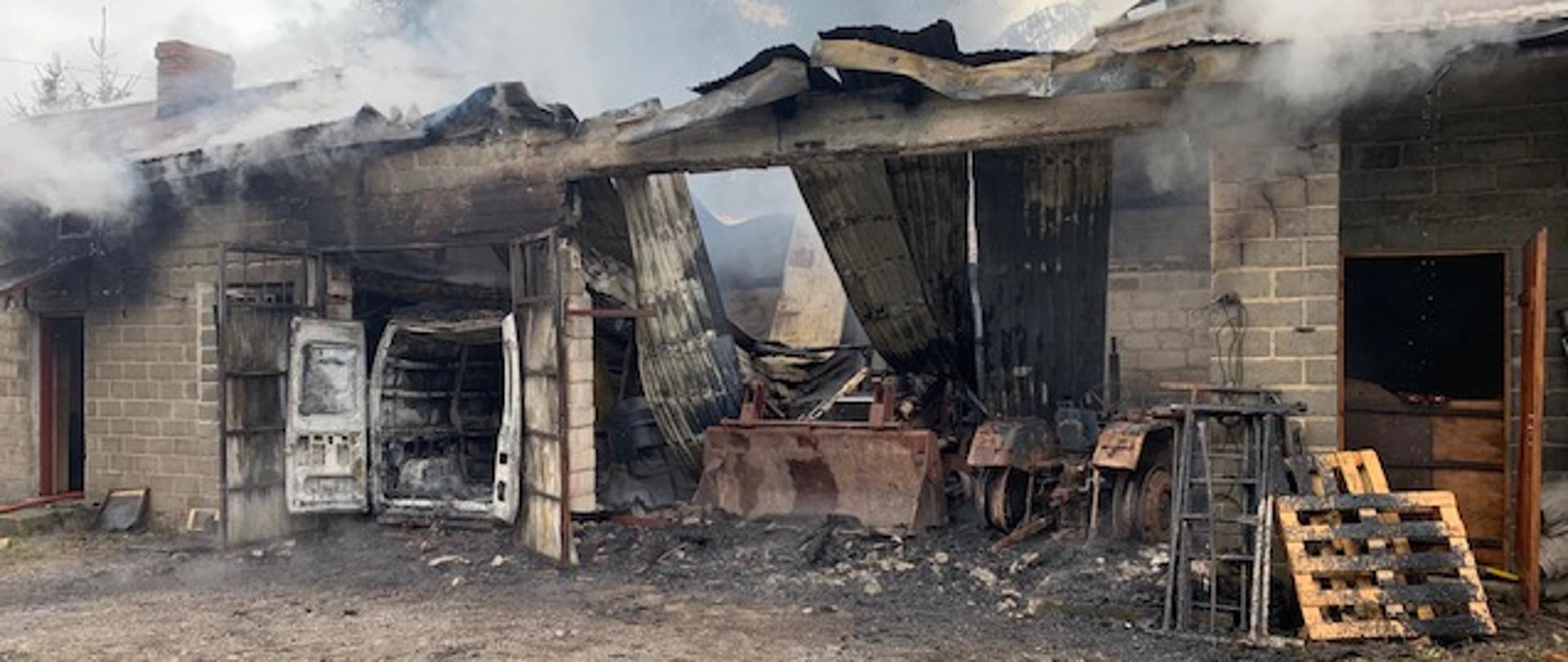 Na zdjęciu widać częściowo spalony budynek gospodarczy, w tle widać spalone pojazdy znajdujące się wewnątrz budynku