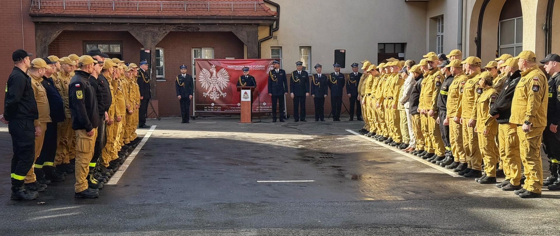 Szerokie ujęcie placu apelowego z góry. Dwa duże pododdziały strażaków stoją naprzeciw siebie, tworząc szeroką aleję. Po lewej stronie widać strażaków w czarnych i piaskowych mundurach, po prawej dominują mundury piaskowe. W głębi, na środku, stoi oficer przy mównicy na tle czerwonej ścianki. Plac jest mokry, odbijają się w nim promienie słońca