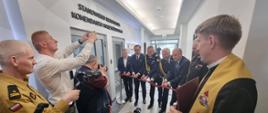 Przedstawiciele mediów stoją pod ścianą, obok stoi kapłan naprzeciwko strażacy Państwowej Straży Pożarnej oraz trzech mężczyzn w garniturach przecinają wstęgę przed wejściem do pomieszczenia nad którym jest napis Stanowisko Kierowania Komendanta Wojewódzkiego.