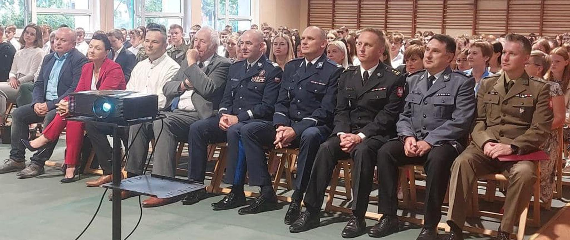 Na zdjęciu znajdują się osoby siedzące na krzesłach ustawione na sali gimnastycznej