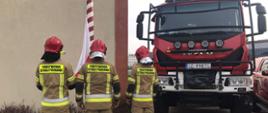 Strażacy Państwowej Straży Pożarnej podnoszą flagę Polski na maszt, obok stoi samochód gaśniczy.