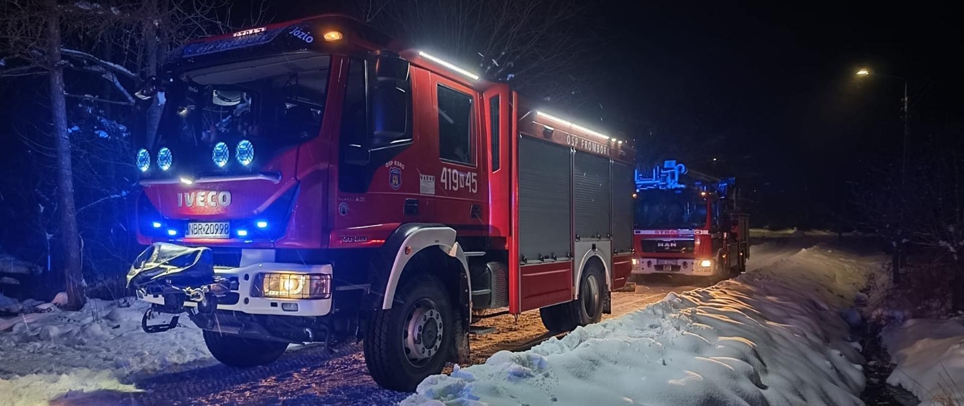 Zima. Noc. Dwa samochody strażackie stoją jeden za drugim z włączonymi światłami.