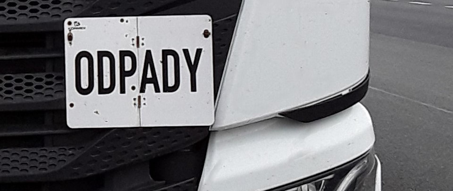 Czarno-biały przód pojazdu z biała tabliczką z czarnym napisem „ODPADY”