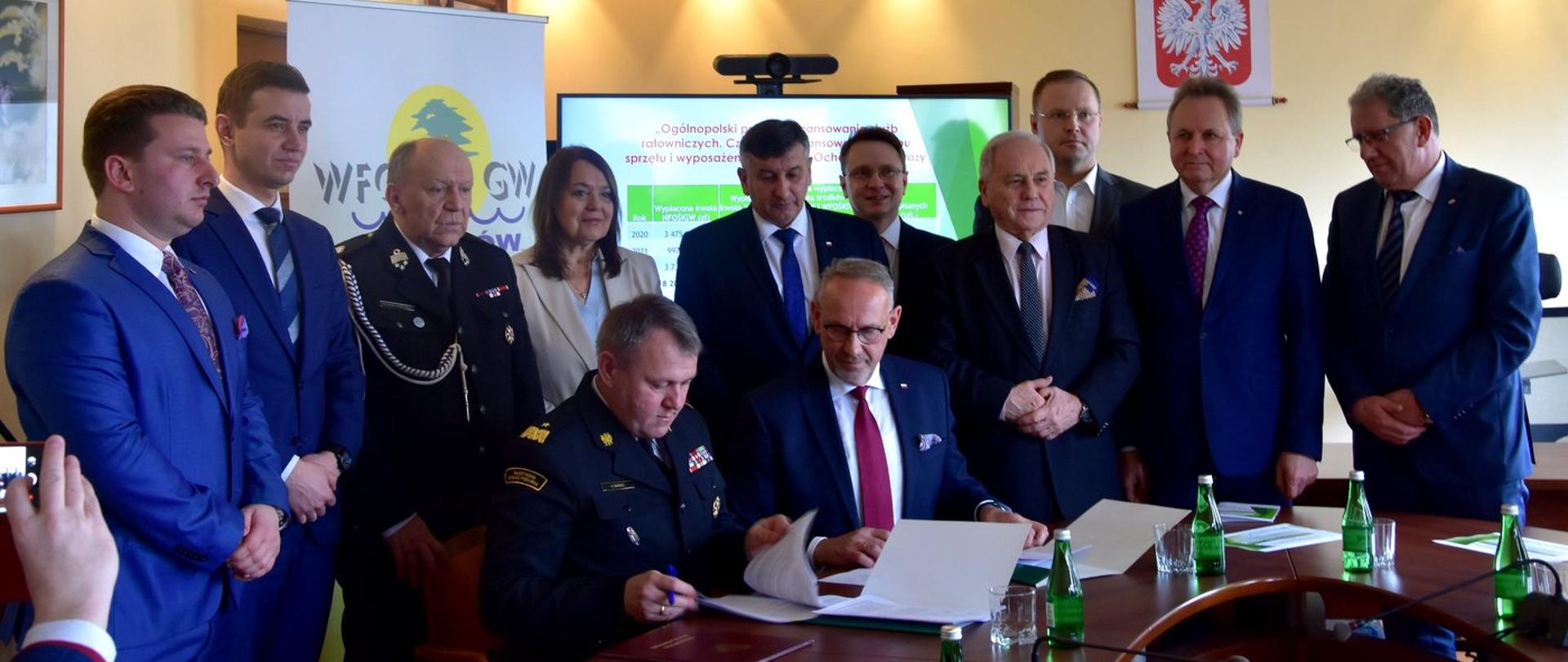 Zdjęcie zrobione w pomieszczeniu. Przy stole siedzi dwóch mężczyzn jeden z nich w mundurze wyjściowym oficera Państwowej Straży Pożarnej. Mężczyźni Podpisują umowy. Za nimi stoją uczestnicy uroczystości