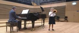 Uczennica stoi przed fortepianem i gra na puzonie, nauczyciel gra na fortepianie na scenie sali koncertowej PSM