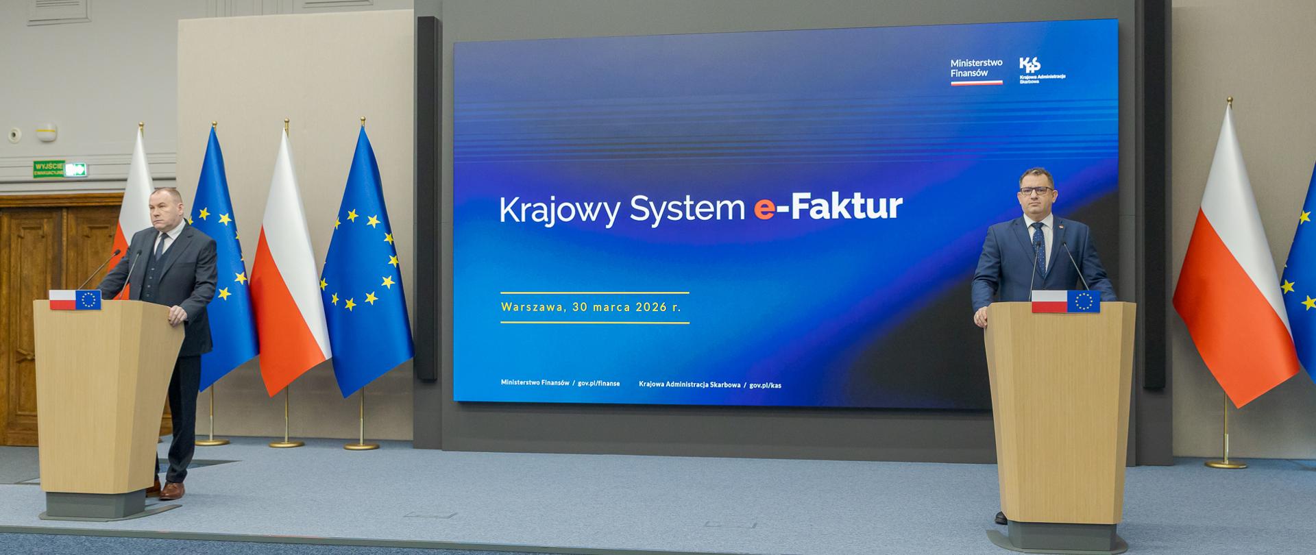 Marcin Łoboda szef Krajowej Administracji Skarbowej oraz Zbigniew Stawicki zastępca szefa KAS podczas konferencji prasowej w Ministerstwie Finansów opowiedzieli o wdrożeniu II etapu Krajowego Systemu e-Faktur przy mównicach przed dużym ekranem z napisem „Krajowy System e‑Faktur”, obok flag Polski i Unii Europejskiej