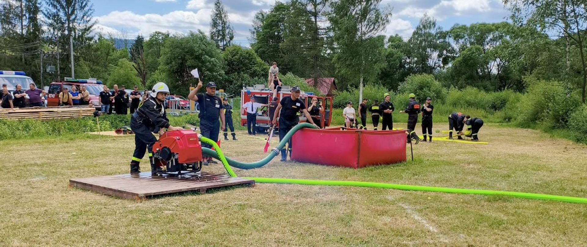 Zdjęcie przedstawia drużynę biorącą udział w zawodach. Na drewnianym podeście stoi strażak przy pompie która zasysa wodę ze zbiornika brezentowego i tłoczy wężami. Widać również sędziów z chorągiewkami - białą i czerwoną. W tle także widać innych strażaków, samochody strażackie i osoby postronne. 