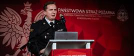 Zastępca komendanta przemawia z mównicy do zaproszonych gości przedstawiając prezentację o historii powstania krajowego systemu ratowniczo-gaśniczego