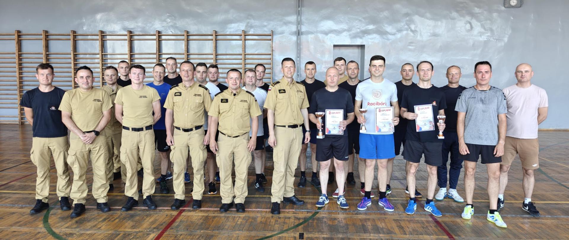 Na zdjęciu znajduje się grupa uczestników i organizatorów turnieju tenisa stołowego pozująca do wspólnej fotografii pamiątkowej w hali sportowej. Wśród zebranych są funkcjonariusze Państwowej Straży Pożarnej w umundurowaniu służbowym oraz uczestnicy zawodów w strojach sportowych. Na pierwszym planie widoczni są laureaci turnieju, którzy prezentują zdobyte puchary i dyplomy.