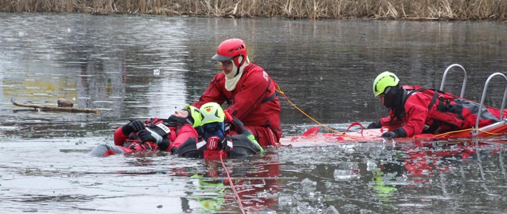 Czterej strażacy w ubraniach do pracy w wodzie ćwiczą na zamarźniętym zbiorniku wodnym ratowanie osoby, pod którą załamał się lód.