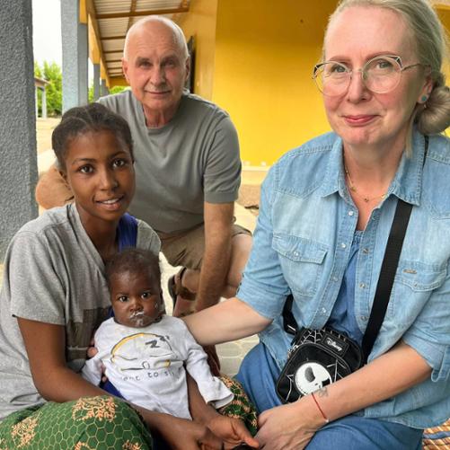 Dwie kobiety trzymają umorusane białą papką niemowlę, za nimi siedzi mężczyzna, z tyłu widać podwórko i kolorowe budynki