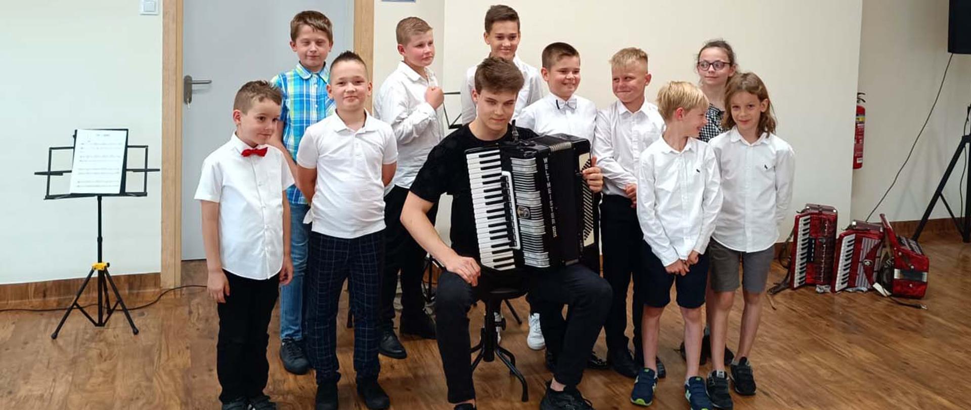 Grupa 10 dzieci stoi zwrócona przodem do robiącego zdjęcie, po środku nich na krześle siedzi starszy chłopiec trzymający na kolanach akordeon. Z tyłu po prawej strony, przy ścianie,na podłodze leży kilka akordeonów.