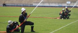 Zdjęcie 2:
Na tym zdjęciu widać kilku strażaków ochotników biorących udział w konkurencji gaśniczej na zielonym boisku. Na pierwszym planie, po lewej stronie, dwóch strażaków klęczy i obsługuje wąż strażacki, z którego pod dużym ciśnieniem wypływa strumień wody, lecący w prawą stronę obrazu. Jeden z nich trzyma dyszę, a drugi pomaga mu stabilizować wąż. Dalej w tle, po prawej stronie, widać kolejne dwie osoby w podobnych pozach, również obsługujące węże. Wszyscy ubrani są w ciemne kombinezony i jasne hełmy strażackie. W tle widać trybuny stadionu i budynki.