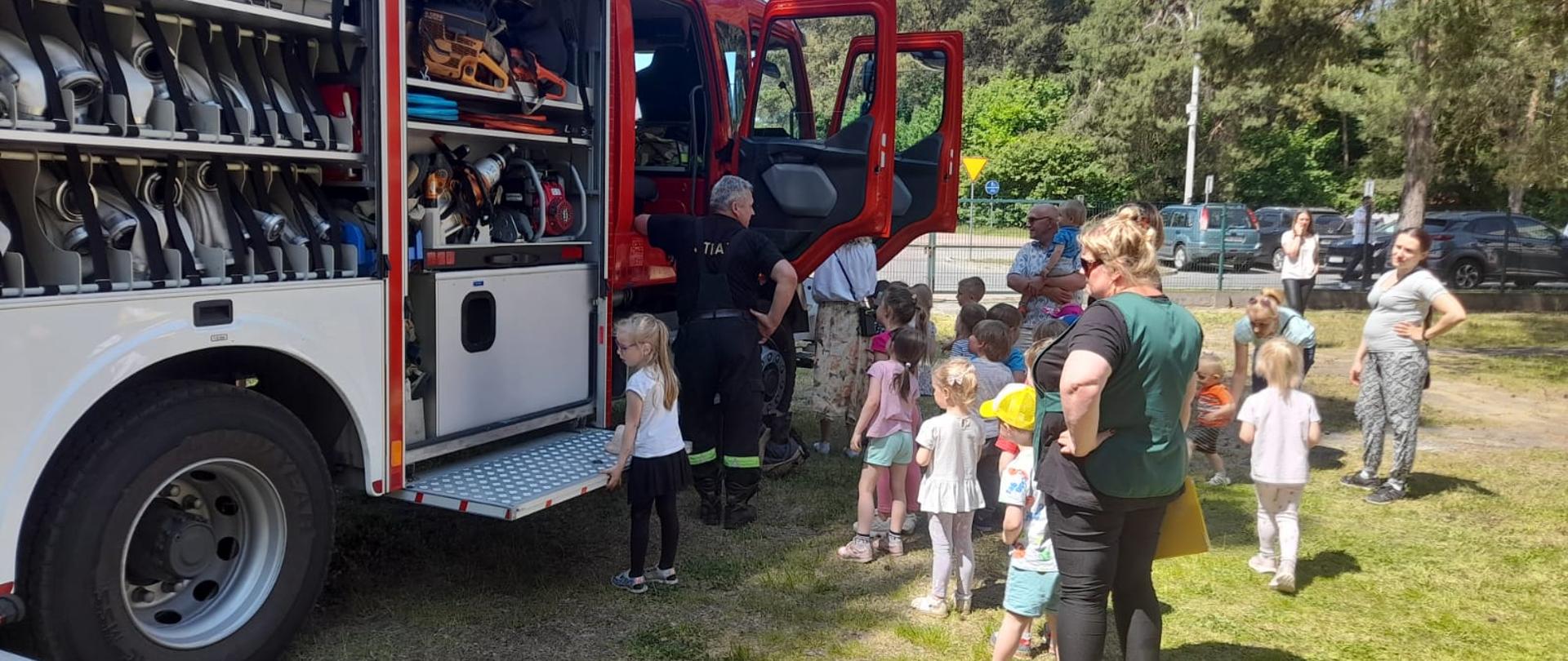 Dzieci z opiekunami stoją przed samochodem pożarniczym w kolejce tak aby każde dziecko mogło wejść do pojazdu i zobaczyć go w środku. W samochodzie w skrytkach widać sprzęt strażacki.
