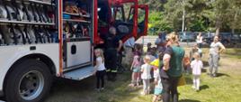 Dzieci z opiekunami stoją przed samochodem pożarniczym w kolejce tak aby każde dziecko mogło wejść do pojazdu i zobaczyć go w środku. W samochodzie w skrytkach widać sprzęt strażacki.