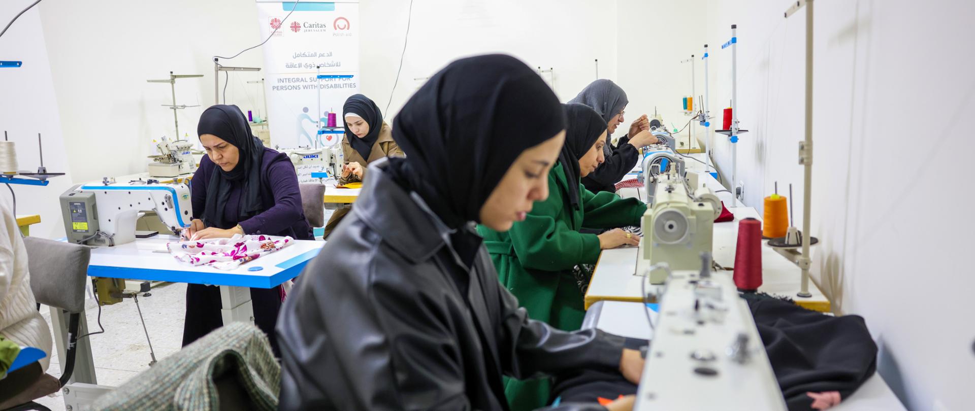 5 kobiet szyje na maszynach, z tyłu widać banner z logo Polska pomoc i Caritas Jerusalem
