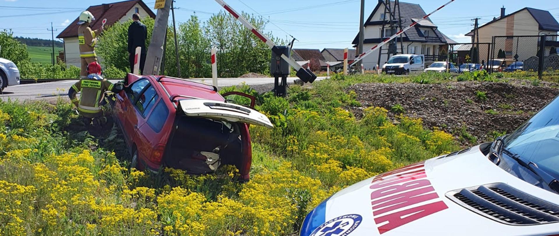 Zdjęcie na pierwszym planie przedstawia przód karetki pogotowia. Na drugim planie znajduje się uszkodzony samochód osobowy oraz betonowy słup oświetleniowy. Po prawej stronie widoczny przejazd kolejowy, na którym zamykają się rogatki. 