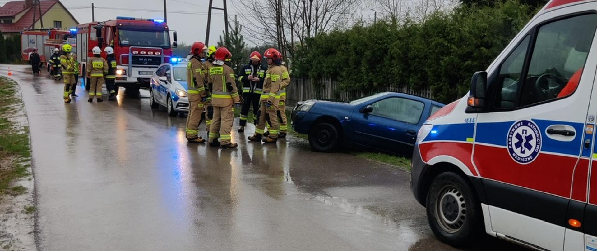 Zdjęcie przedstawia stojący w rowie uszkodzony samochód osobowy. Na jezdni stoją zaparkowane pojazdy służb ratowniczych oraz strażacy.