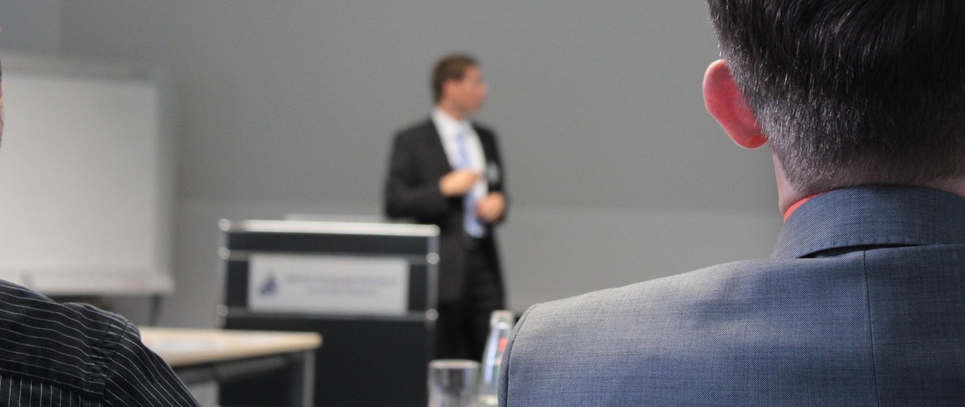 Szkolenie - w tle widoczny prowadzący, na pierwszym planie tyłem mężczyzna w garniturze
