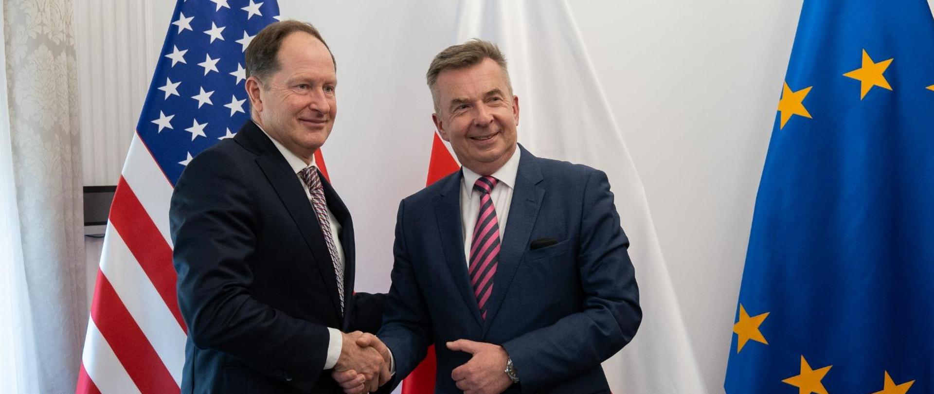 Minister Wieczorek stoi obok mężczyzny w garniturze, podają sobie ręce, za nimi pod białą ścianą flagi Polski, UE i USA.