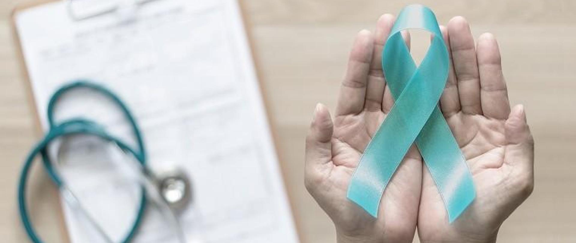 Na zdjęciu przysunięte do siebie otwarte dłonie na których leży zielona wstążeczka po lewej stronie w tle stetoskop leżący na biurku