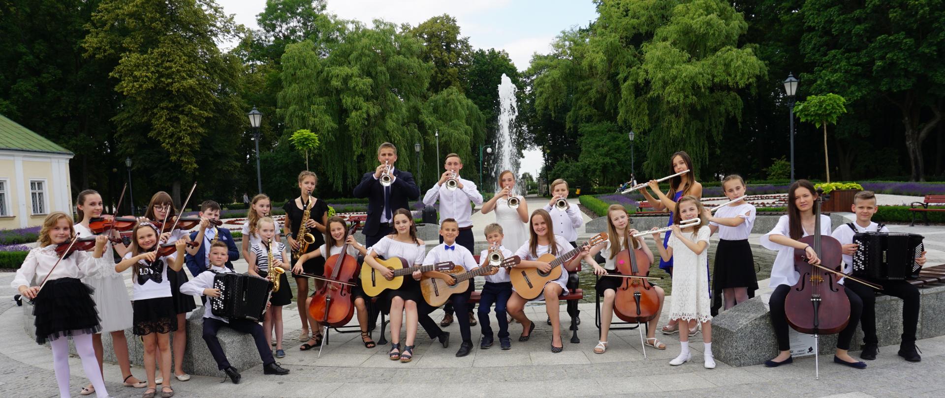 Uczniowie PSM I stopnia w Końskich ubrani na galowo z instrumentami w rękach pozują do zdjęcia w parku. Z tyłu za nimi fontanna.